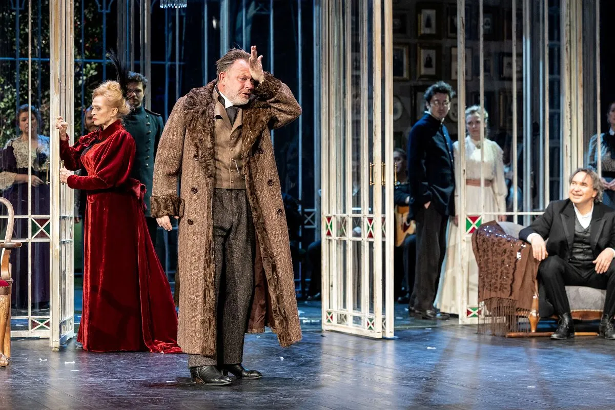 Aktor w brązowym płaszczu z futrzanym kołnierzem gestykuluje na scenie. Obok niego kobieta w czerwonej sukni. To scena z 