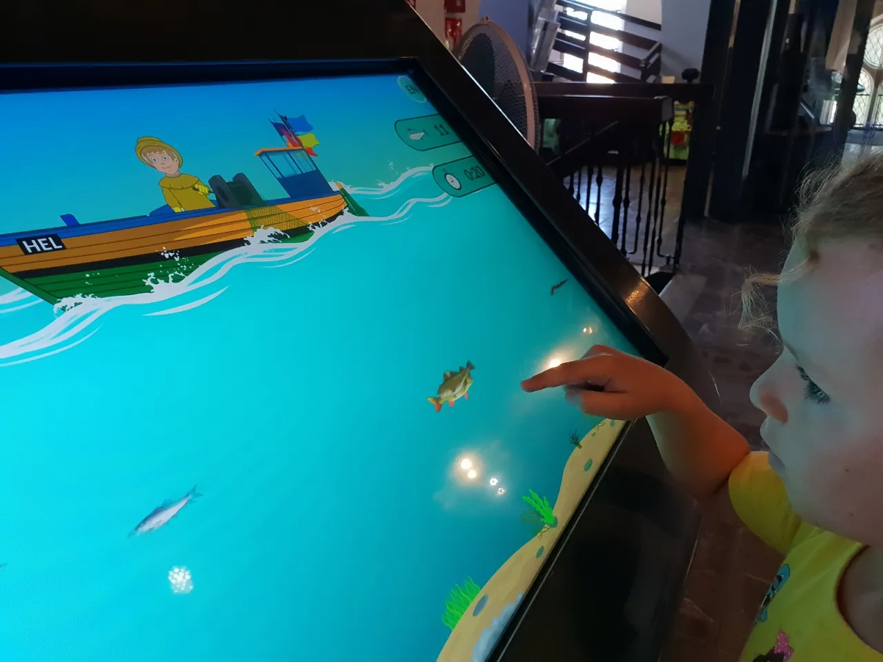 Dziecko wskazuje palcem na rybę na interaktywnym ekranie, gdzie łódź z napisem