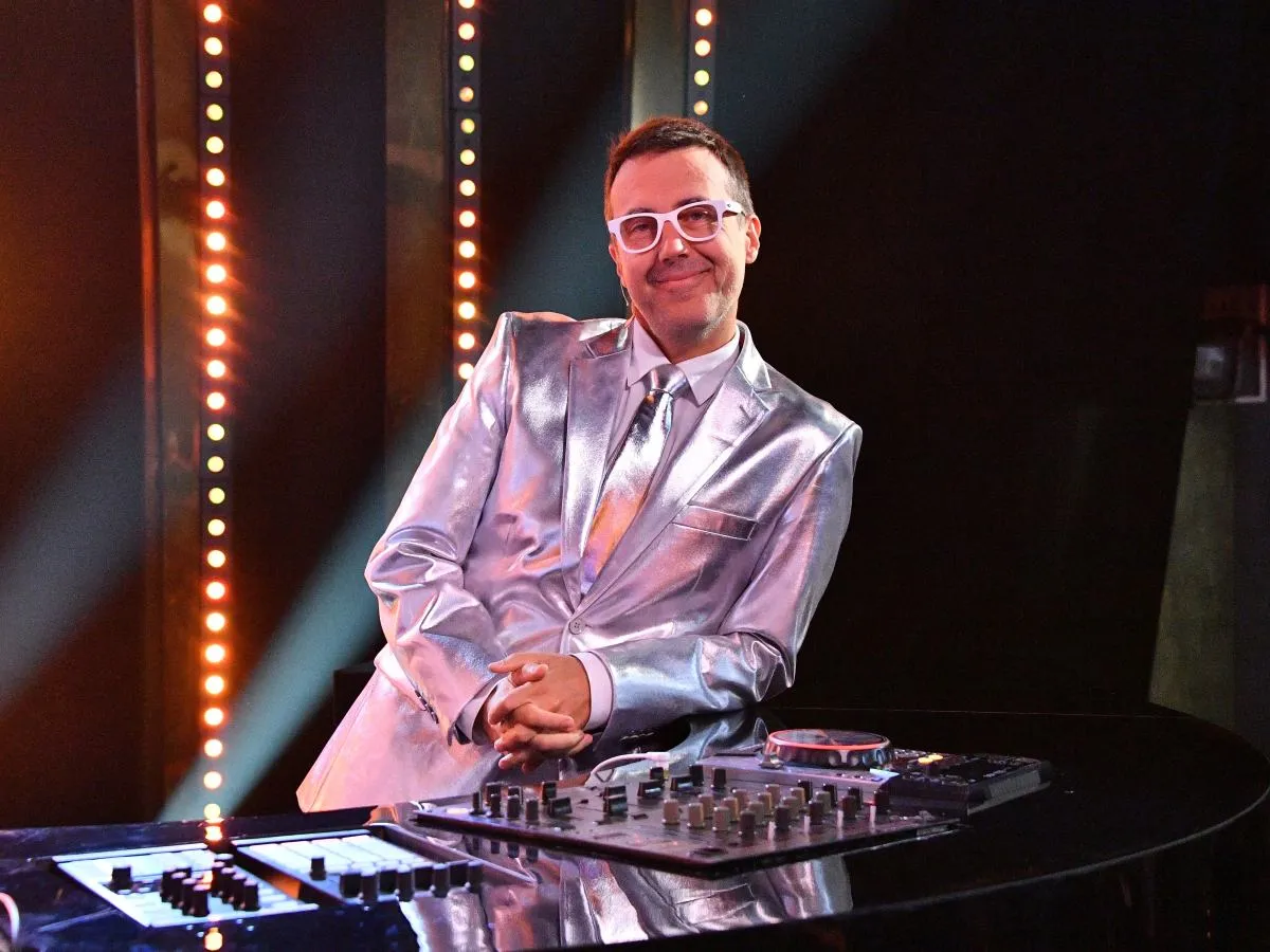 DJ Adamus w srebrnym garniturze, z białymi okularami, przy konsoli DJ-skiej. Jaka to melodia?