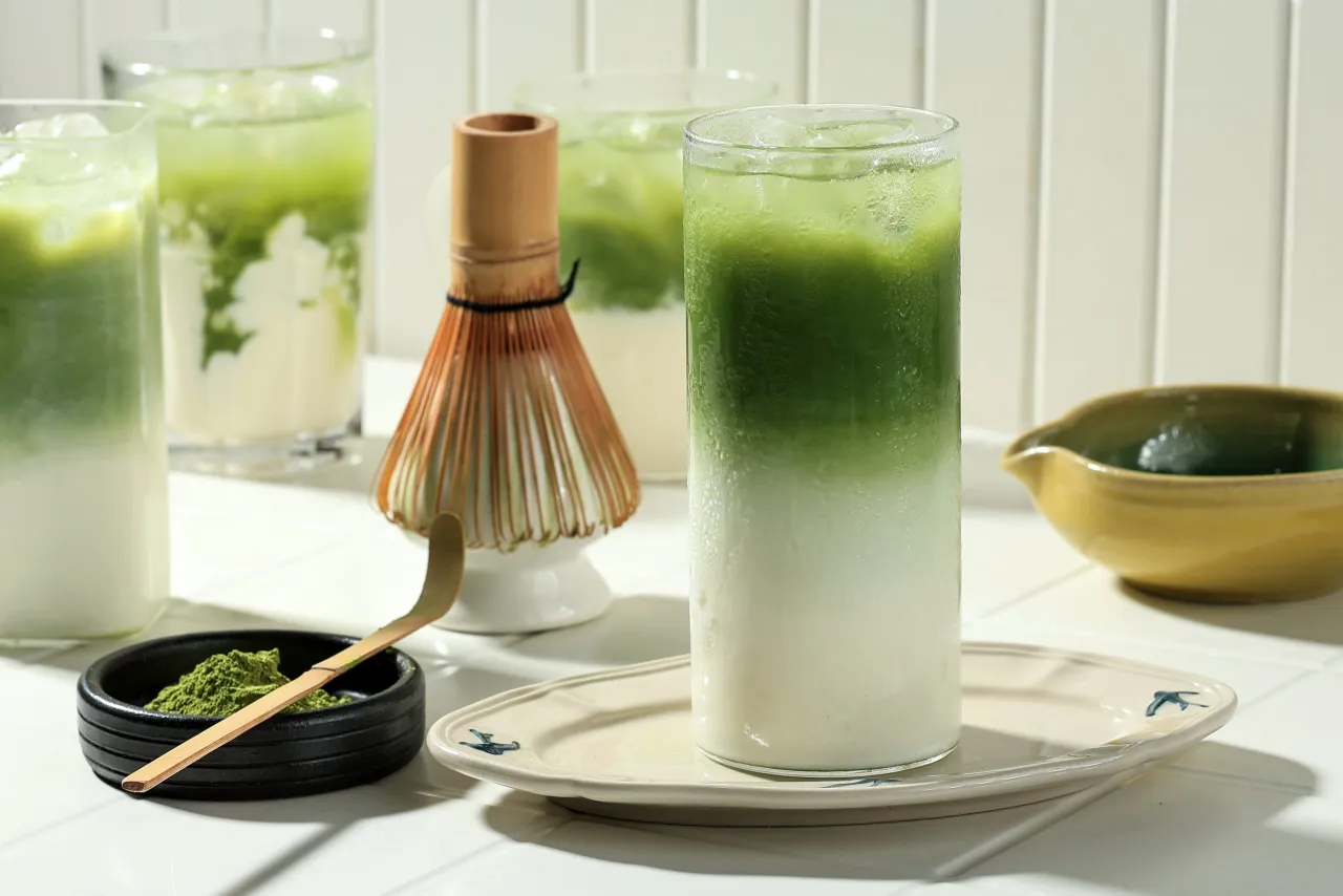 Orzeźwiający nap&oacute;j z matchy z lodem i mlekiem, idealny na co dzień. Obok bambusowa miotełka i proszek matcha.