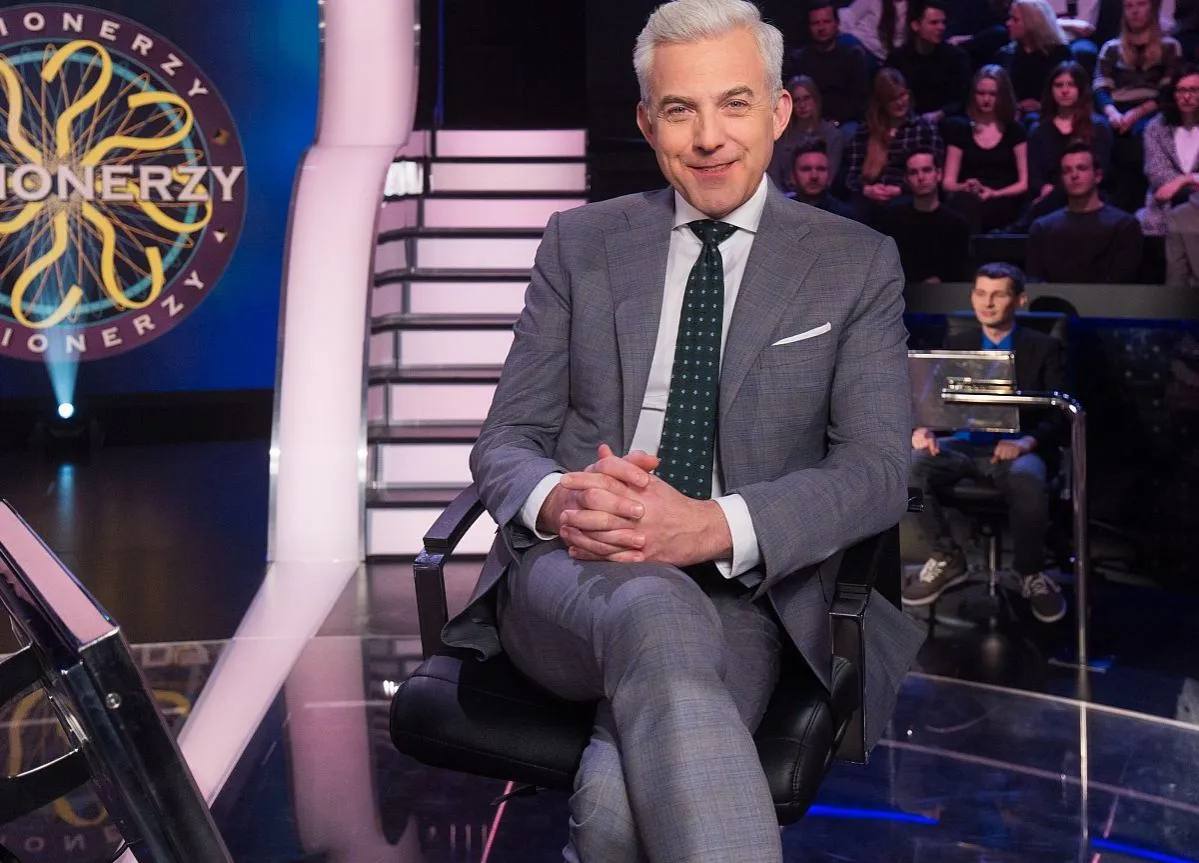 Uśmiechnięty prowadzący w garniturze siedzi na scenie teleturnieju 