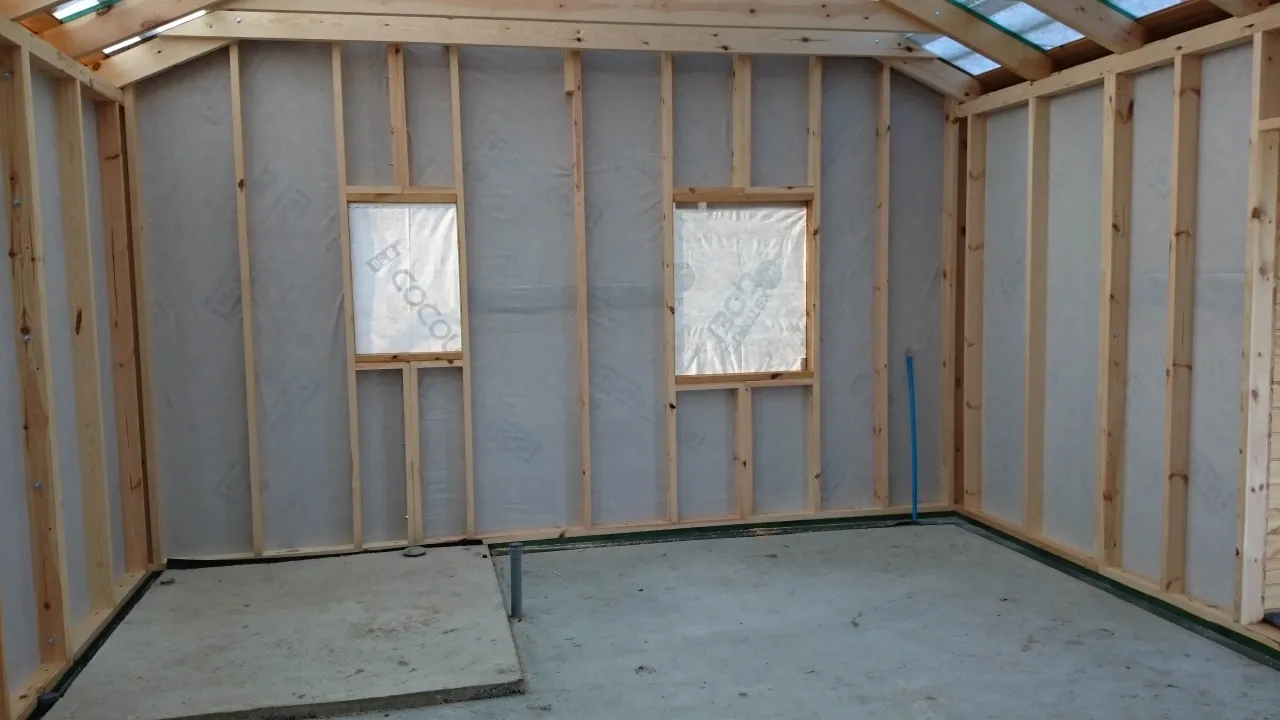 Wnętrze domku drewnianego w budowie. Widoczne szkieletowe ściany, okna, dach i betonowa podłoga. To początek prac, jak zrobić konstrukcje domku drewnianego.