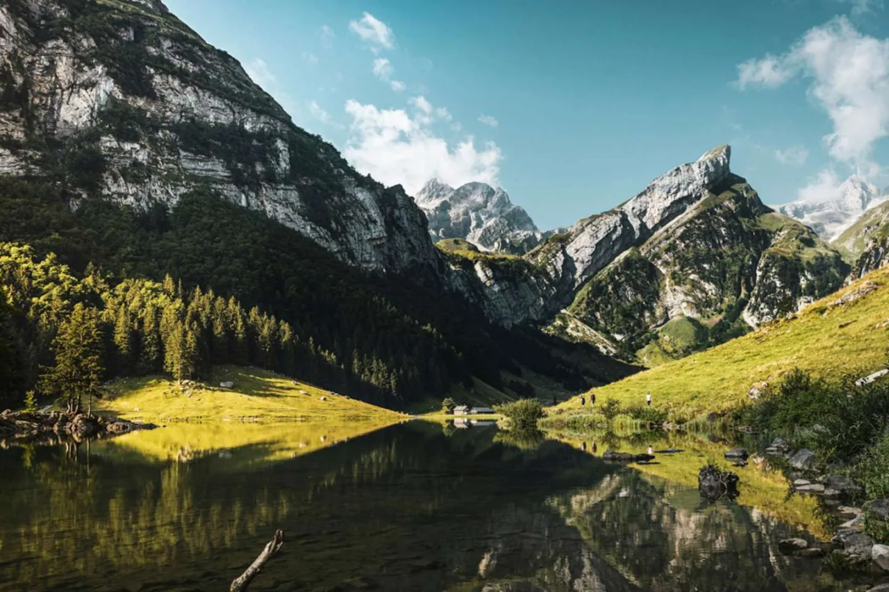 Szwajcaria latem: spokojne jezioro odbija skaliste szczyty i zielone zbocza g&oacute;r.