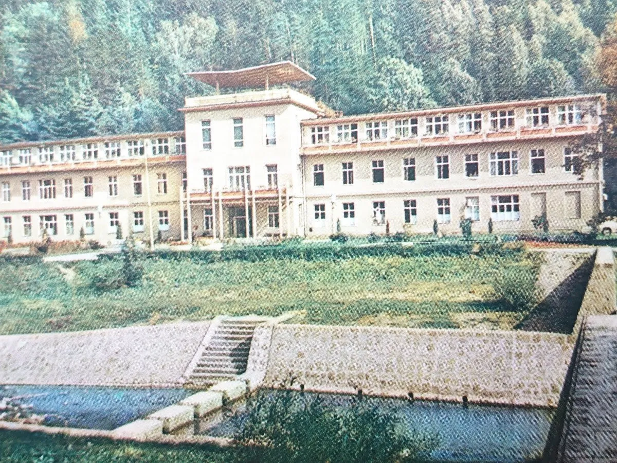 Sanatoria w Rymanowie Zdroju, rozległy budynek z tarasem na dachu, otoczony zielenią i strumieniem.