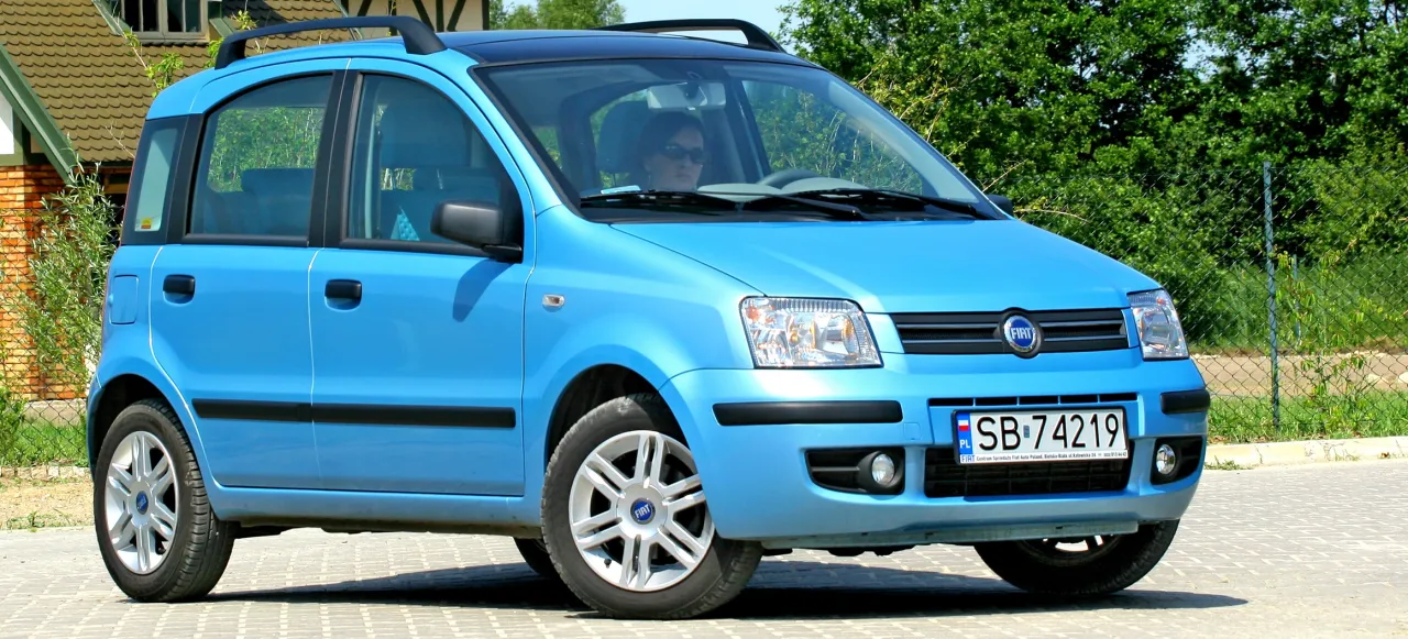 Błękitny Fiat Panda, pierwszy samoch&oacute;d z panoramicznym dachem, zaparkowany na kostce brukowej.