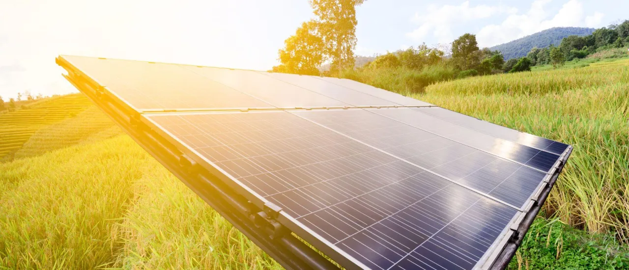 Panele fotowoltaiczne na tle zielonych p&oacute;l i g&oacute;r. Słońce oświetla instalację, sugerując korzyści z inwestycji w fotowoltaikę.