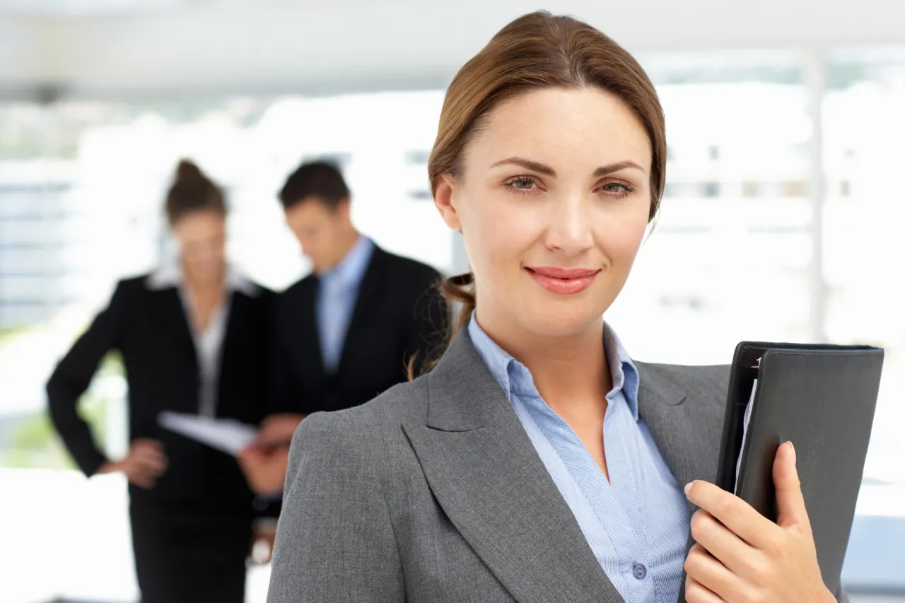 Uśmiechnięta bizneswoman z teczką, w tle wsp&oacute;łpracownicy omawiający dokumenty. Widać, że panuje między nimi umowa dorozumiana.