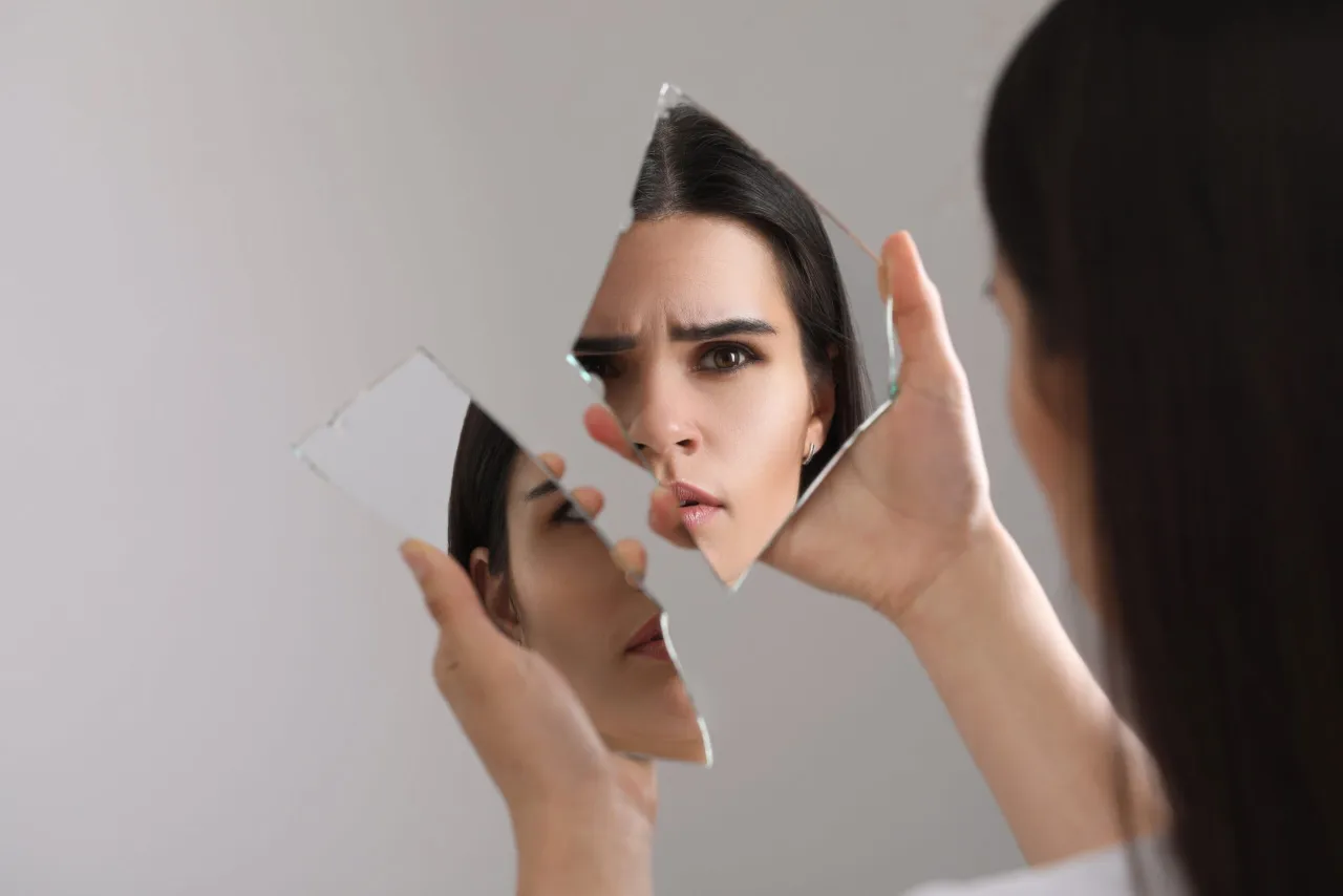 Kobieta patrzy na swoje odbicie w połamanej tafli lustra, zastanawiając się, czym są kompleksy i jak wpływają na postrzeganie siebie.