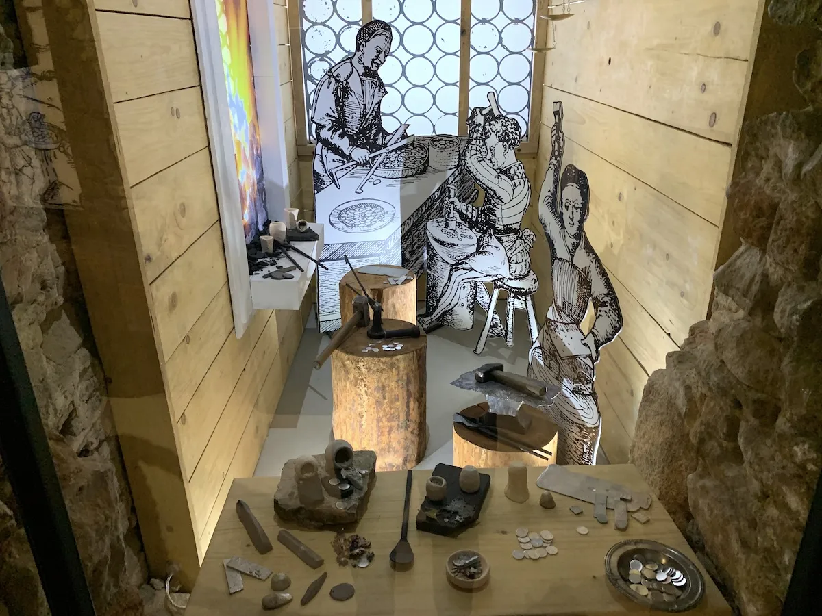 Scena z legendy o Olkuszu: rzemieślnicy pracują przy stołach z narzędziami i monetami.