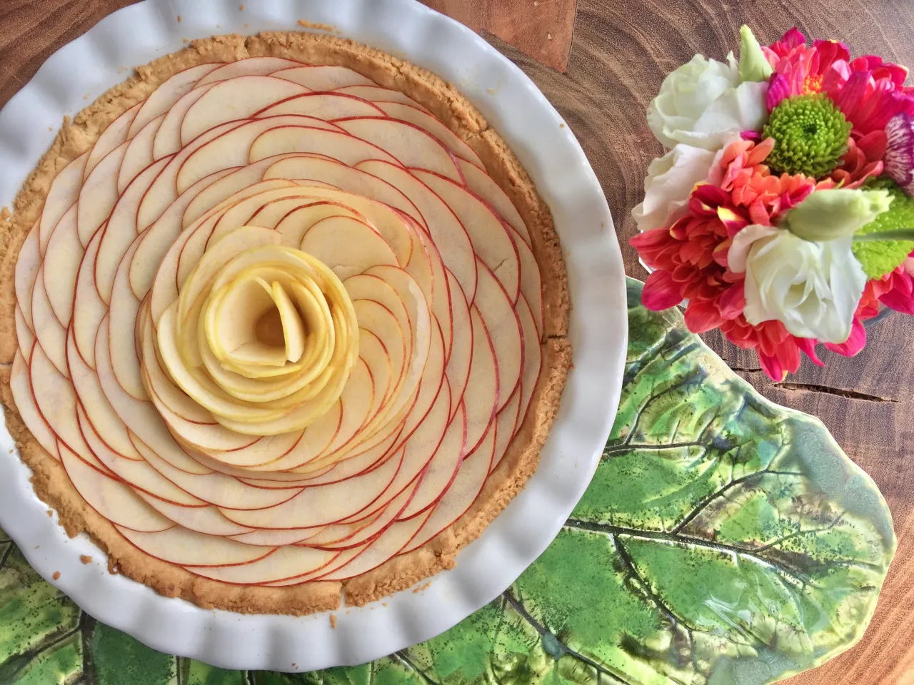 Przepis na kruche ciasto z jabłkami ułożonymi w kształt r&oacute;ży. Idealne na deser, obok bukiet kolorowych kwiat&oacute;w.