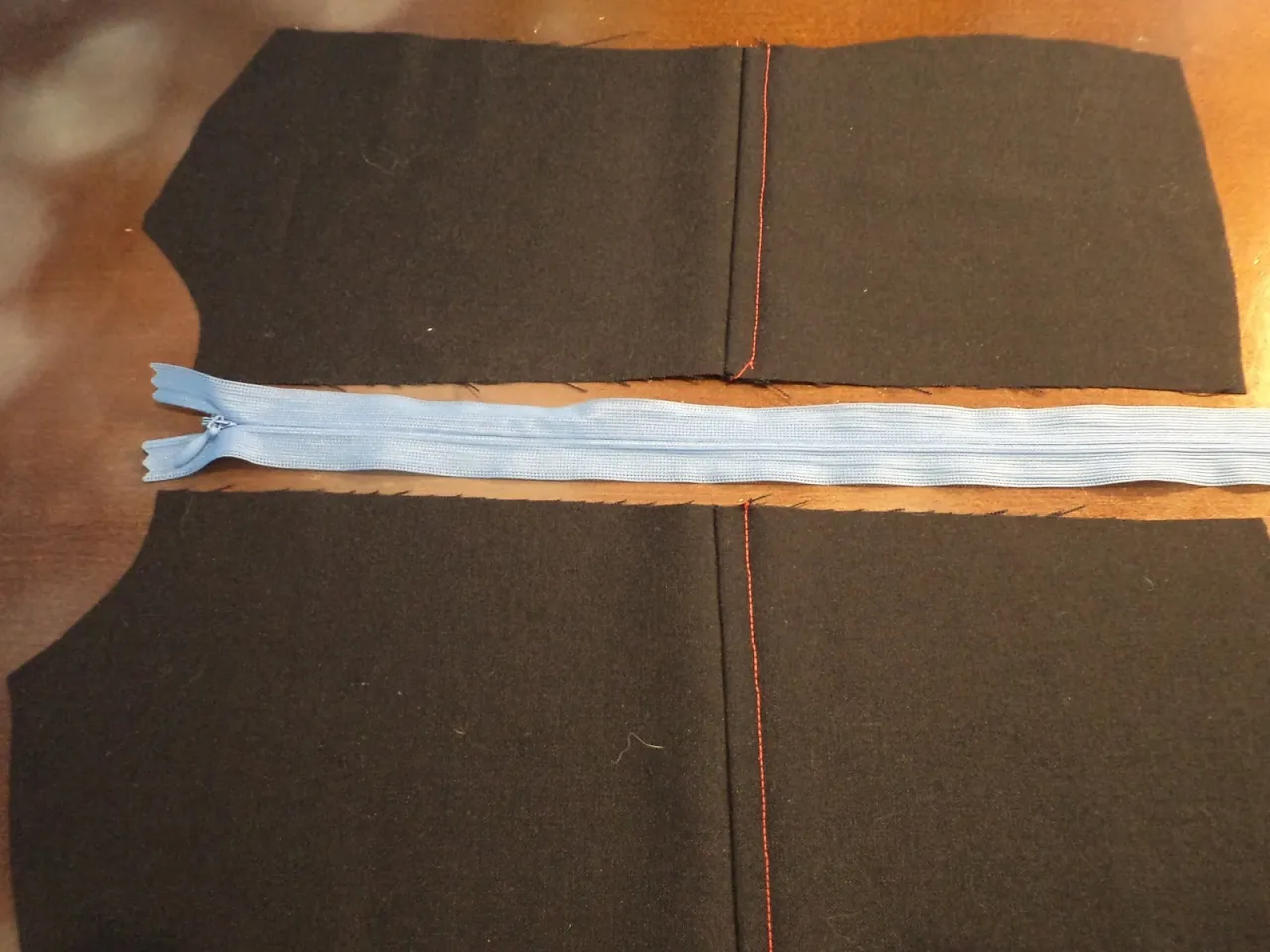 Przygotowanie do wszywania suwaka. Dwa kawałki czarnego materiału z czerwonymi szwami po bokach, z jasnoniebieskim suwakiem ułożonym między nimi.