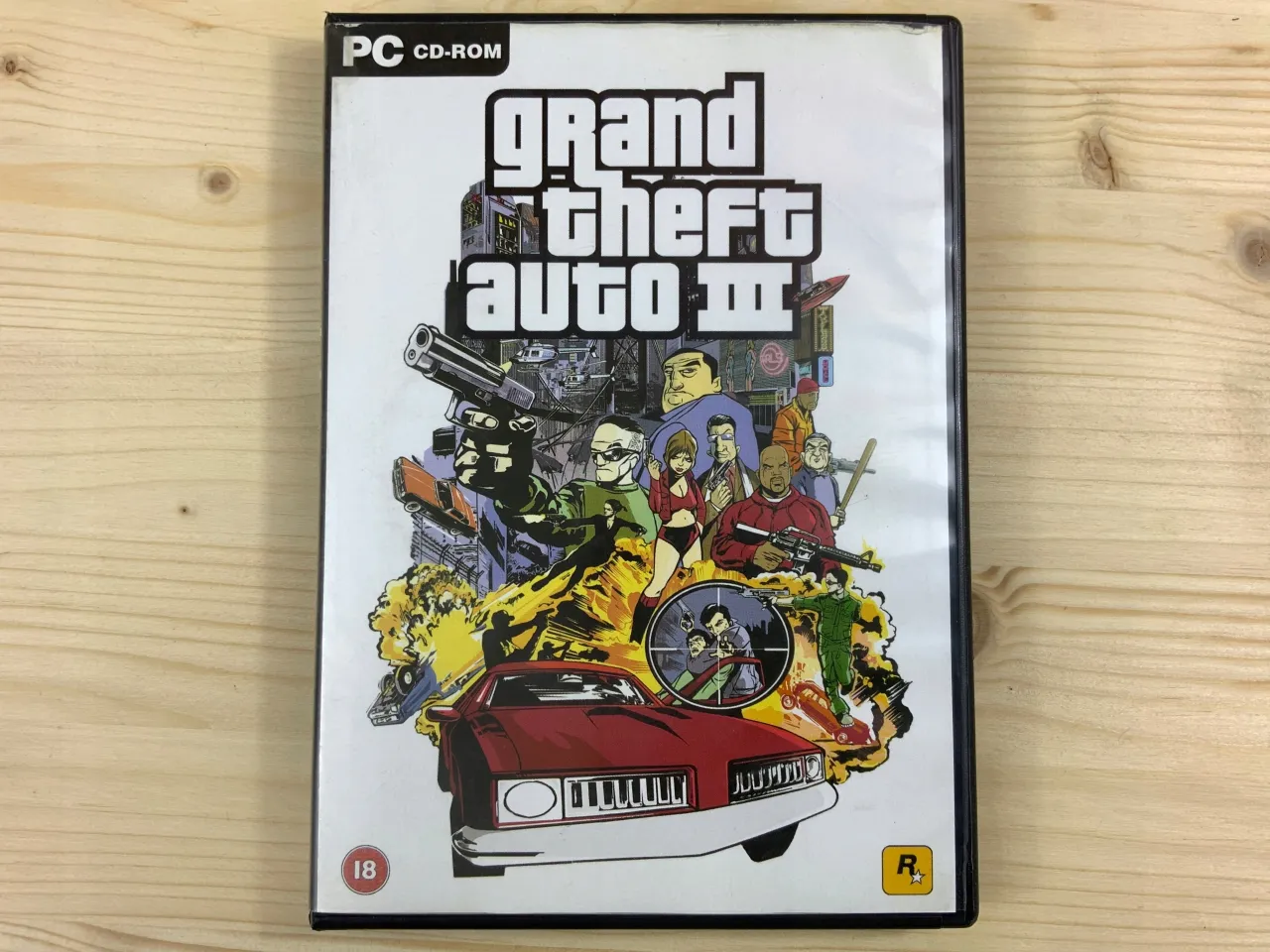 Okładka gry Grand Theft Auto III na PC. W tle dynamiczna scena z postaciami i wybuchami, a na pierwszym planie czerwony samochód. To idealne miejsce na gta 3 spolszczenie!