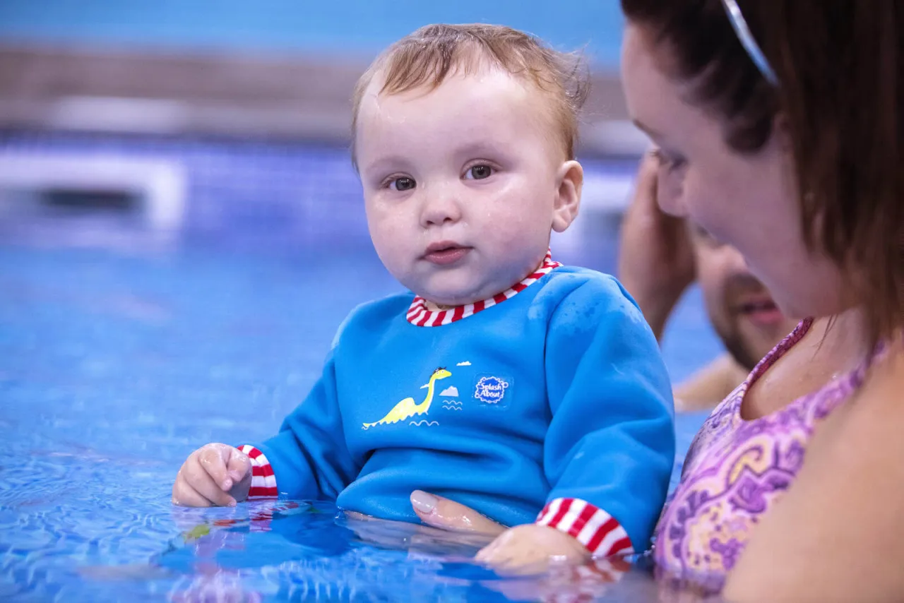 Niebieski kombinezon do pływania dla dzieci z ż&oacute;łtym dinozaurem, z czerwono-białymi mankietami i kołnierzykiem, w wodzie z mamą.
