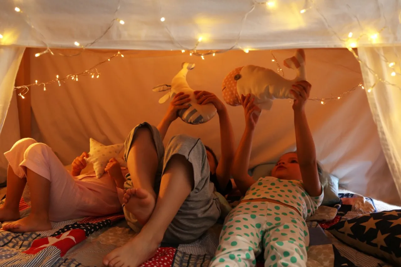 Dzieci w domku z kocy i lampek, bawią się pluszowymi wielorybami. Idealny pomysł, jak zrobić bazę w domu.