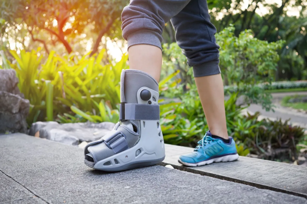 Osoba w bucie ortopedycznym i sportowym bucie stawia pierwsze kroki po artroskopii stawu skokowego. Długość rehabilitacji jest indywidualna.