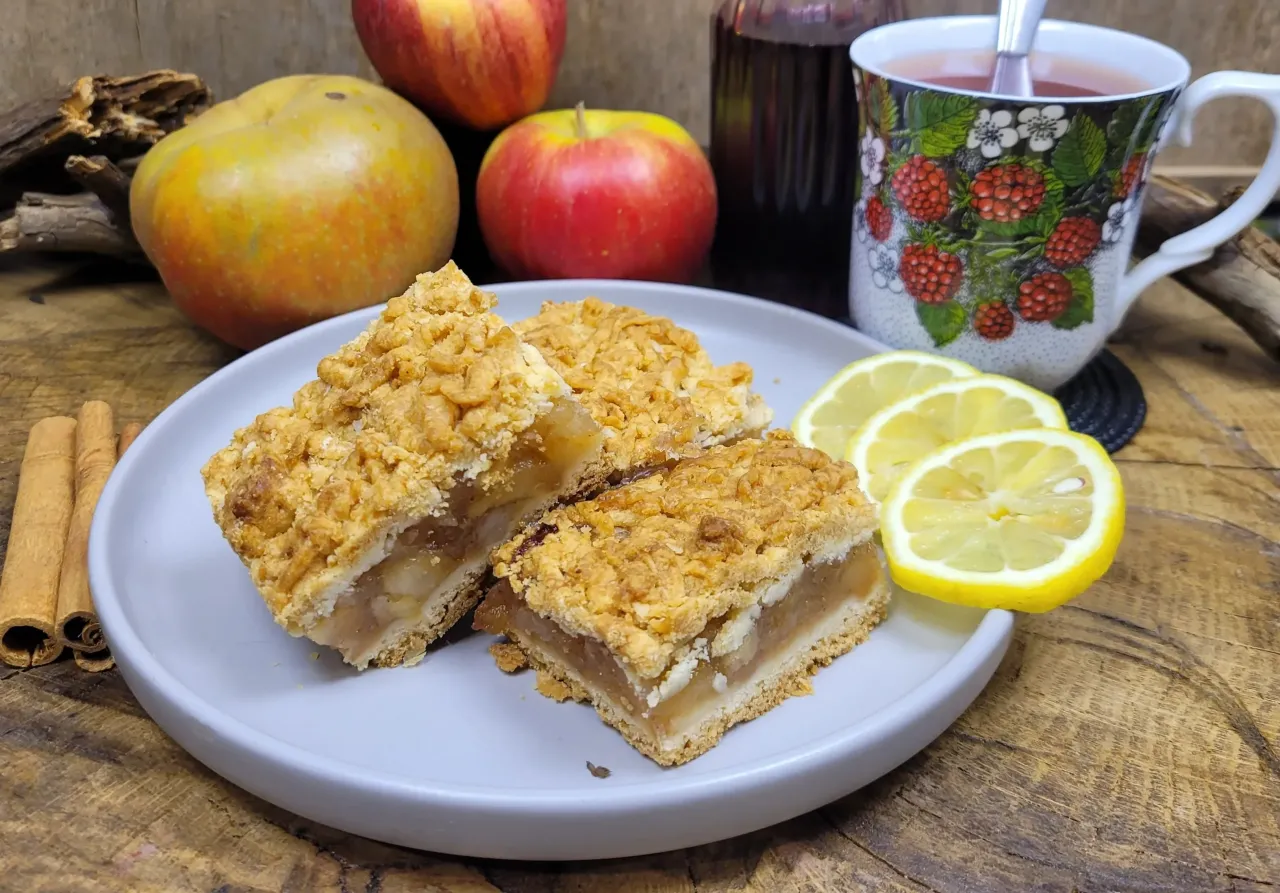Kawałki pysznej szarlotki na talerzu, obok jabłka, cytryny i cynamonu. Idealne na prosty przepis na szarlotkę.