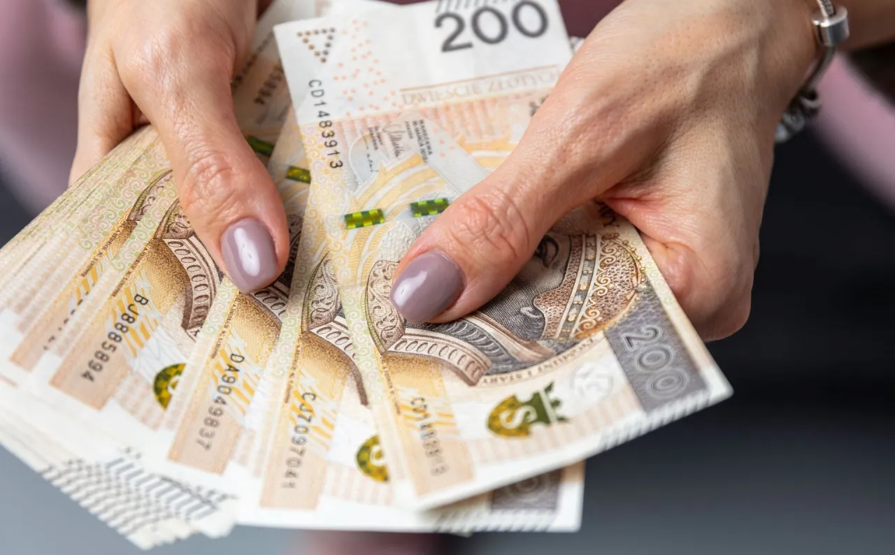 Dłonie trzymające stos banknotów 200 zł. Zastanawiasz się, jak długo wypłacana jest okresowa emerytura kapitałowa?