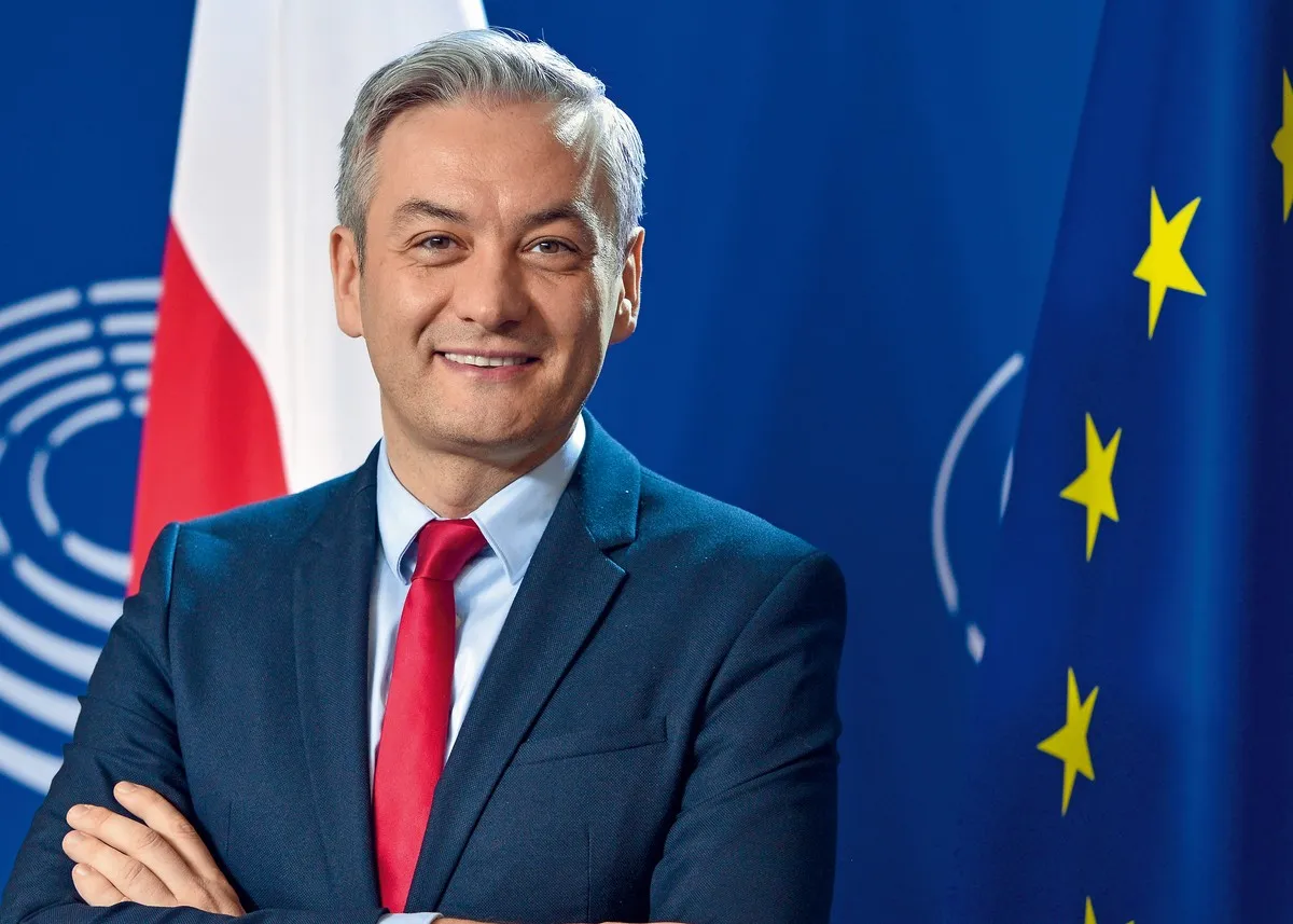 Robert Biedroń, polityk, uśmiecha się na tle flagi Polski i UE. Ile lat ma Robert Biedroń?