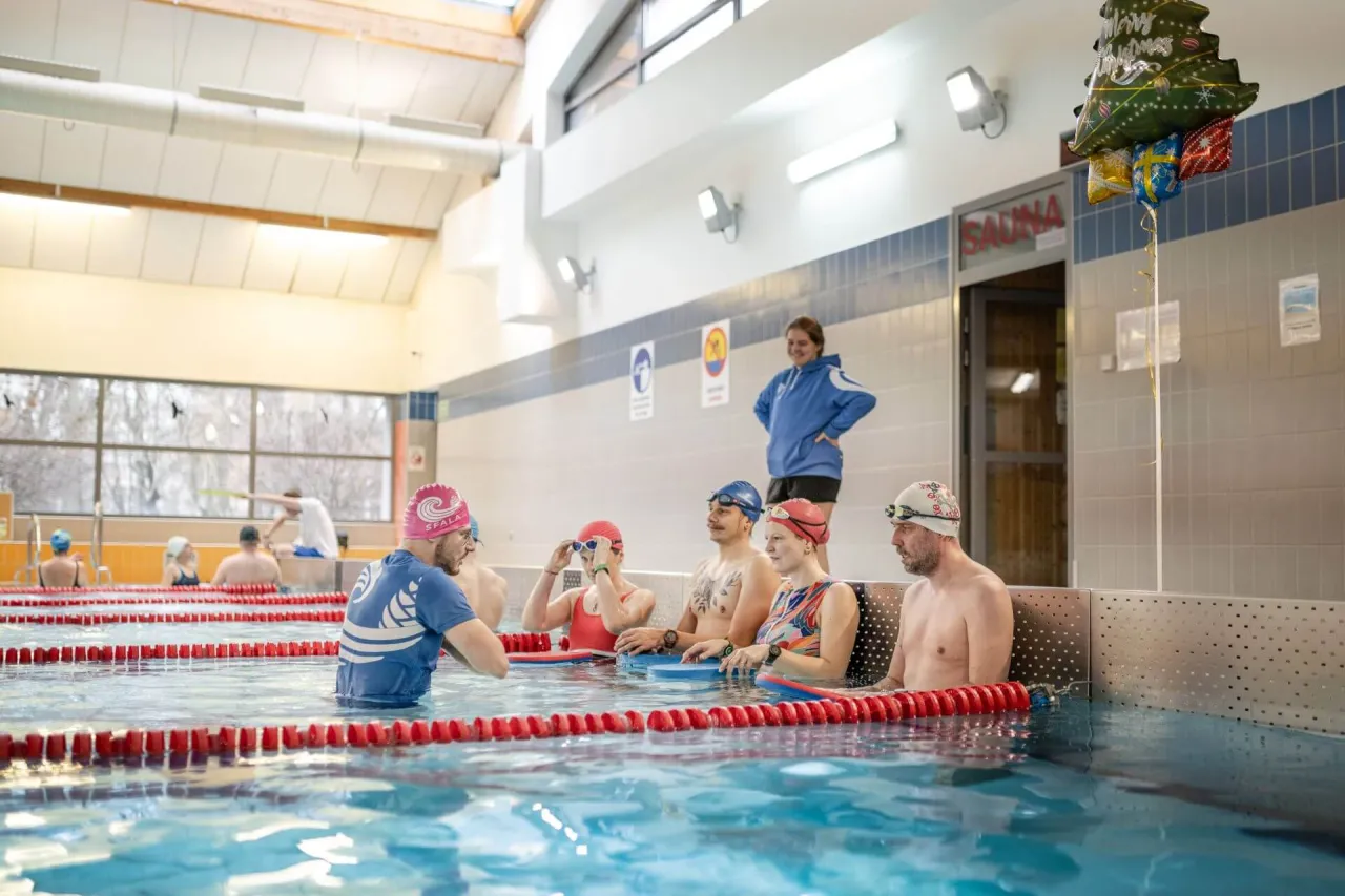 Instruktor wyjaśnia, jak nauczyć pływać dorosłego. Grupa osób w czepkach i okularach pływackich słucha uważnie w basenie.