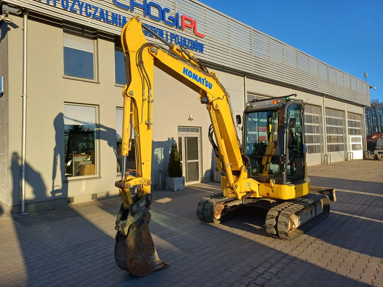Żółta koparka Komatsu czeka na swoją godzinę pracy przed budynkiem z napisem 