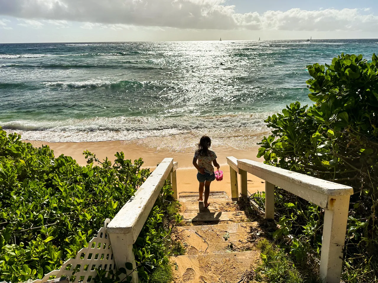 Dziewczynka schodzi po schodach na plażę, gdzie leżą Hawaje na mapie. Słońce odbija się od fal oceanu.