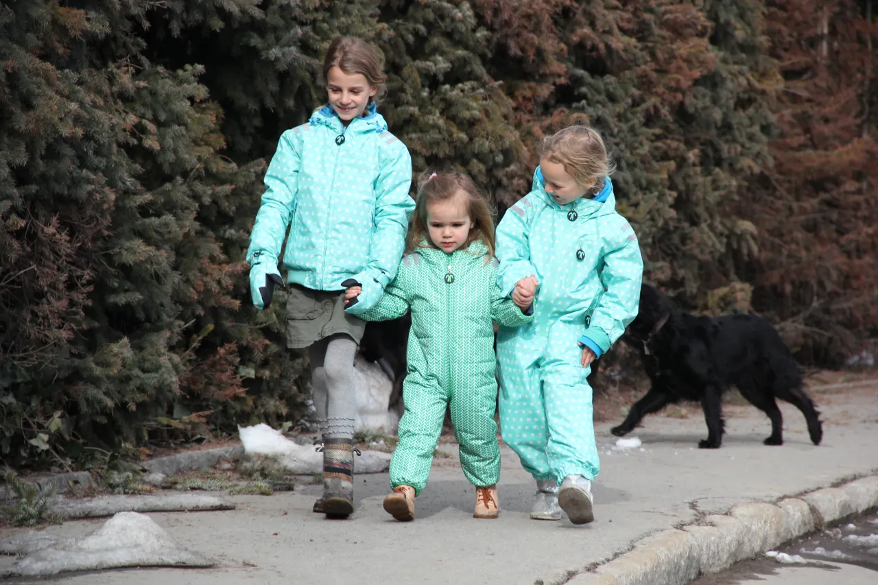 Trzy dziewczynki w zimowych kombinezonach i kurtkach spacerują z psem.