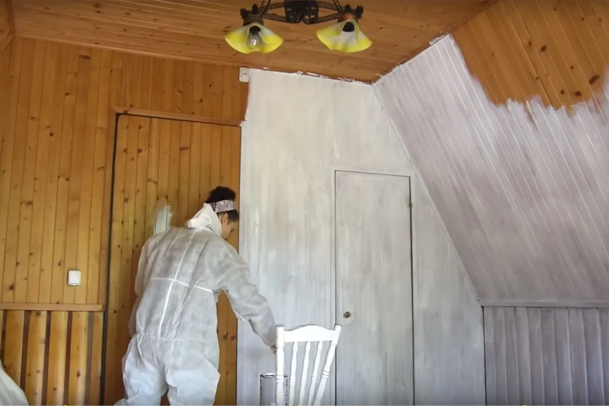 Kobieta w kombinezonie maluje boazerię na biało. Widać postęp prac, jak pomalować boazerię.