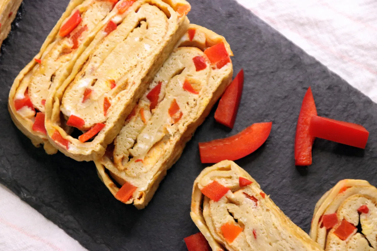 Rolowany omlet tamago z kawałkami czerwonej papryki, ułożony na czarnej desce.