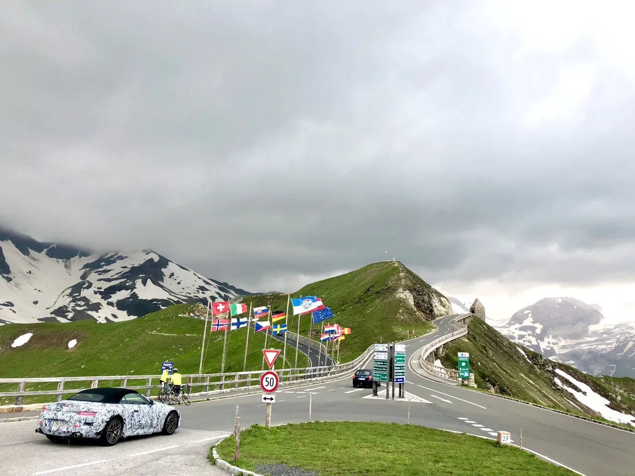 Wjazd na g&oacute;rską serpentynę, jedną z najpiękniejszych dr&oacute;g w Europie, z widokiem na ośnieżone szczyty i flagi państw.