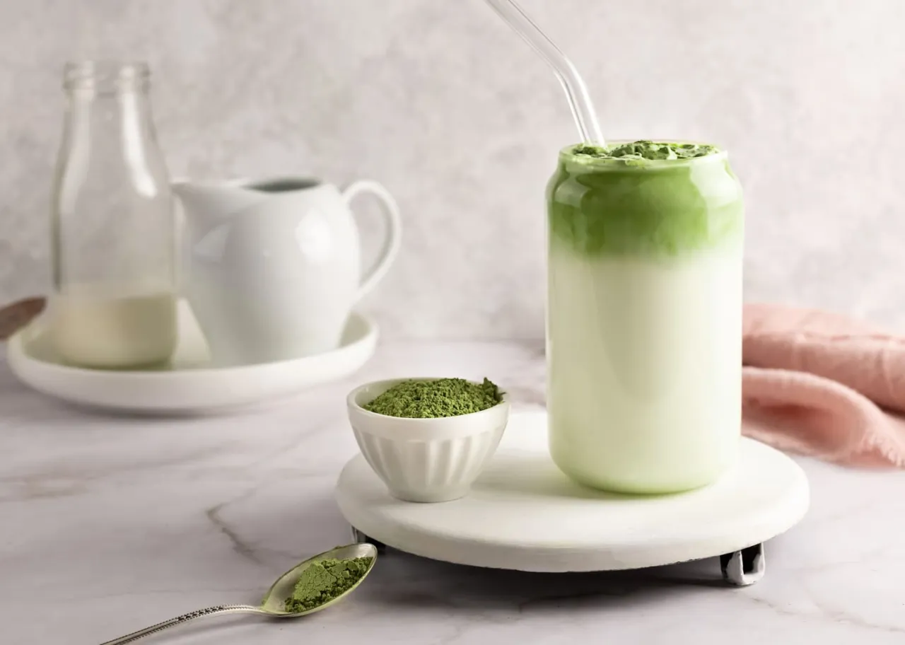 Zielona herbata matcha w szklance z mlekiem, obok miseczka z proszkiem i łyżeczka. Idealny sposób, jak pić matcha latte.