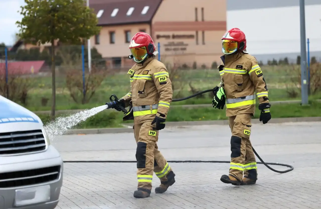 Dwóch strażaków w akcji. Jeden z nich kieruje strumień wody na pojazd. Ciekawe, ile zarabia strażak zawodowy za taką pracę.