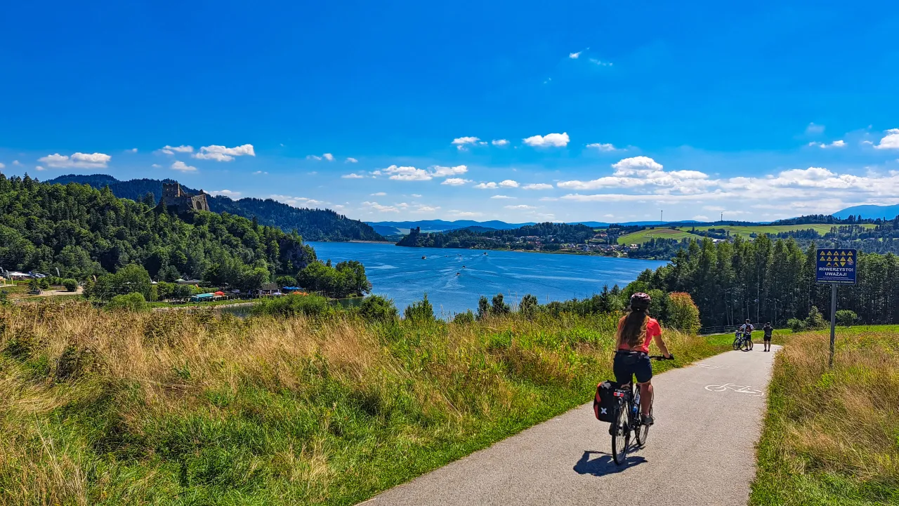 Kobieta na rowerze przemierza malowniczą trasę rowerową wokół jeziora, podziwiając ruiny zamku na wzgórzu i błękitne niebo.
