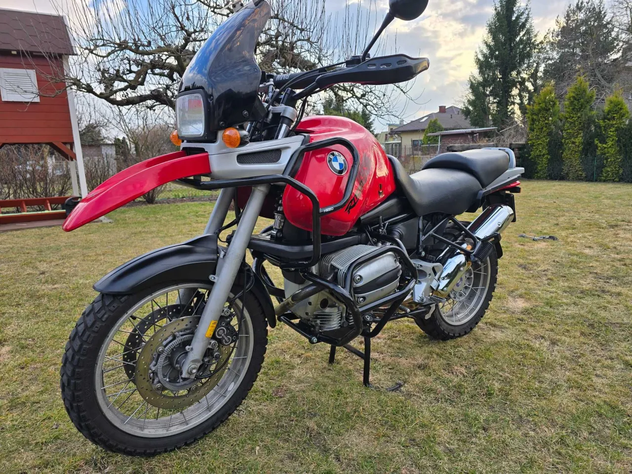 Czerwony motocykl BMW R 1100 R z czarną szybą i kuframi, zaparkowany na trawie w ogrodzie.