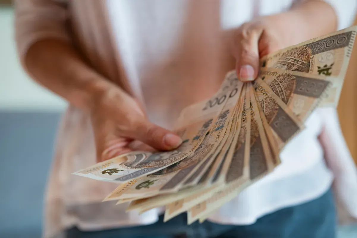 Dłonie trzymają wachlarz banknotów 200 zł. Zastanawiasz się, czy nagroda jubileuszowa wlicza się do emerytury?