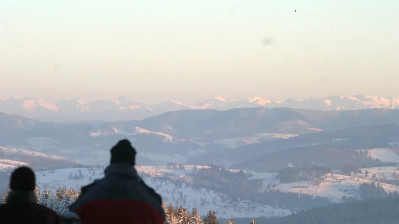 Dwie osoby patrzą na ośnieżone g&oacute;ry. W oddali widać pasmo Tatr, a poniżej łagodne wzg&oacute;rza Beskid&oacute;w.