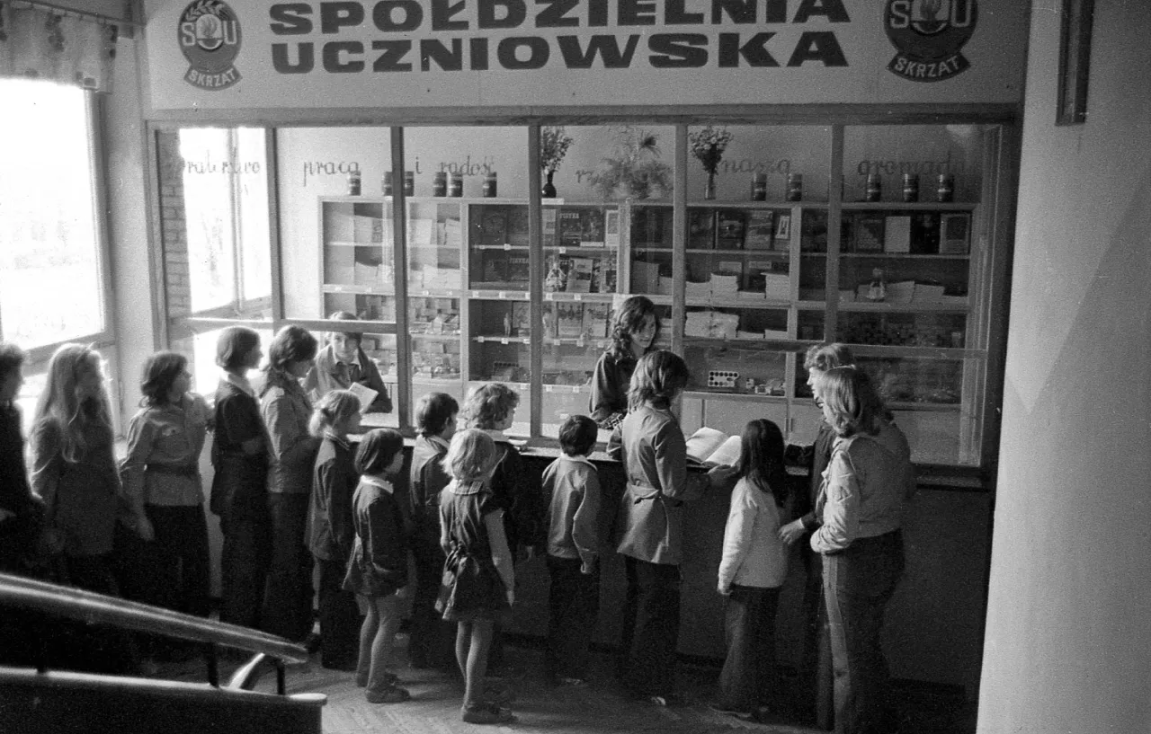 Dzieci stoją w kolejce do sklepiku szkolnego, gdzie można kupić zeszyty, książki i inne artykuły.