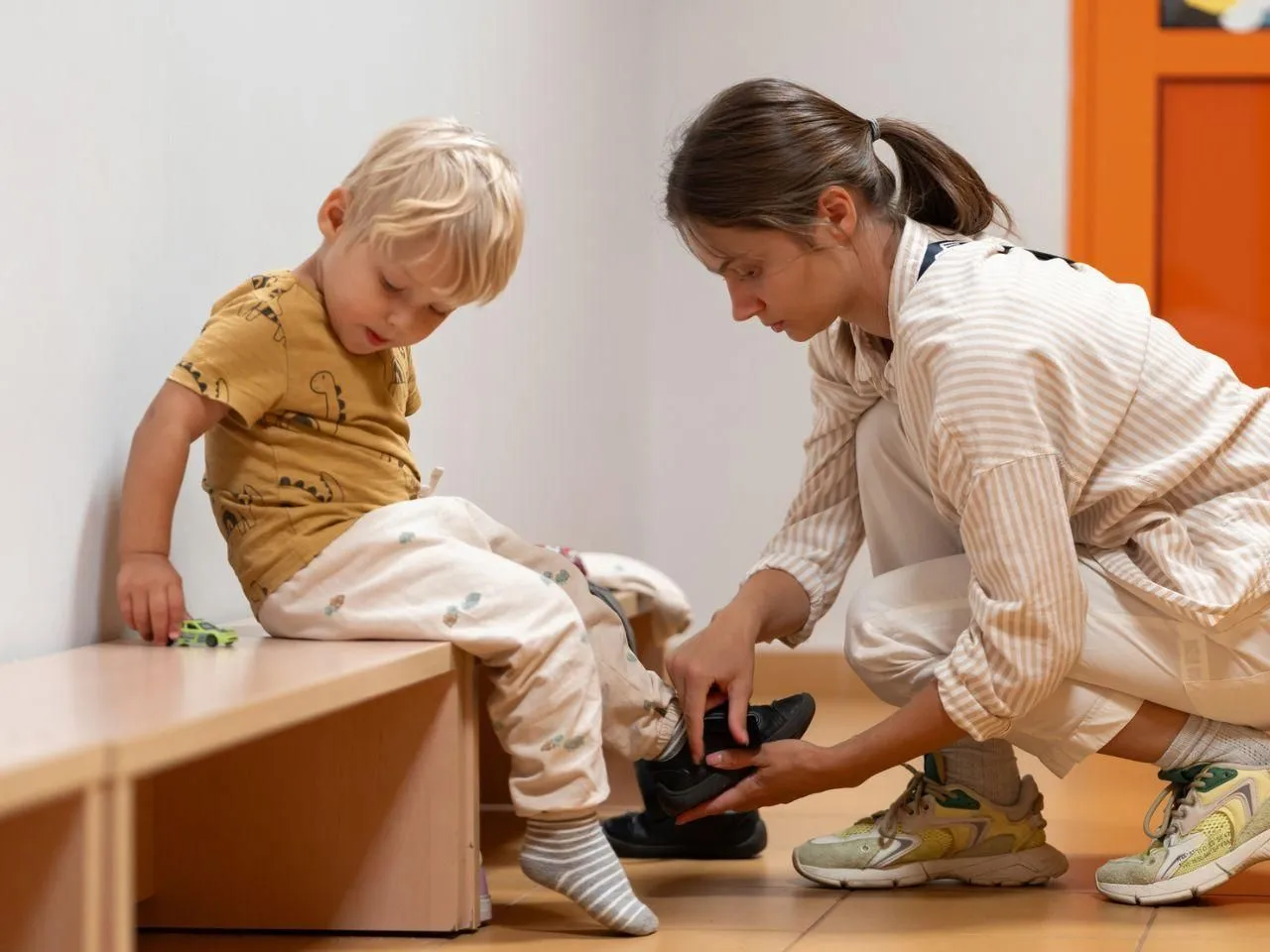 Dziecko 5 lat z blond włosami siedzi na ławce, a kobieta pomaga mu założyć buty.