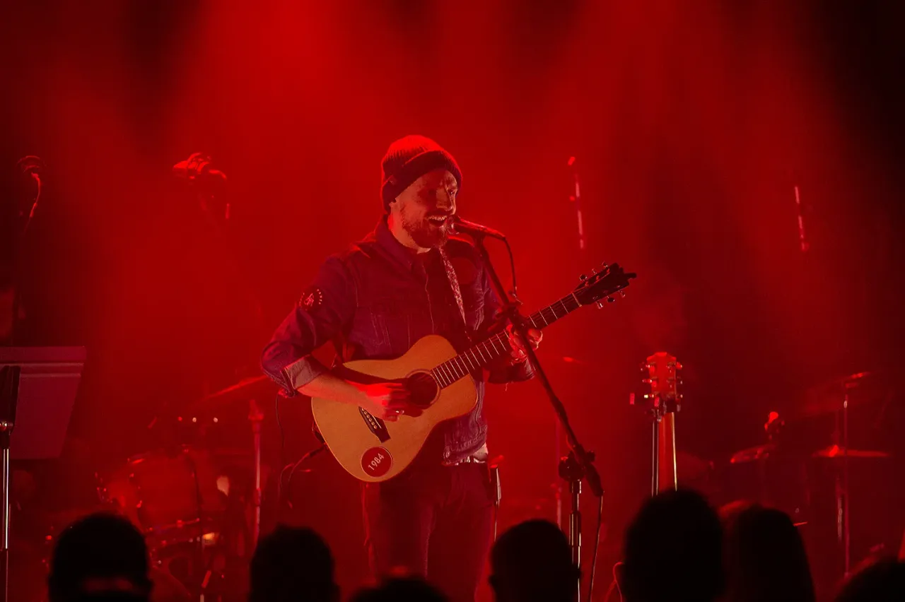 Artysta z gitarą na scenie, w czerwonym świetle. Fani zastanawiają się, ile trwa koncert Domagały.