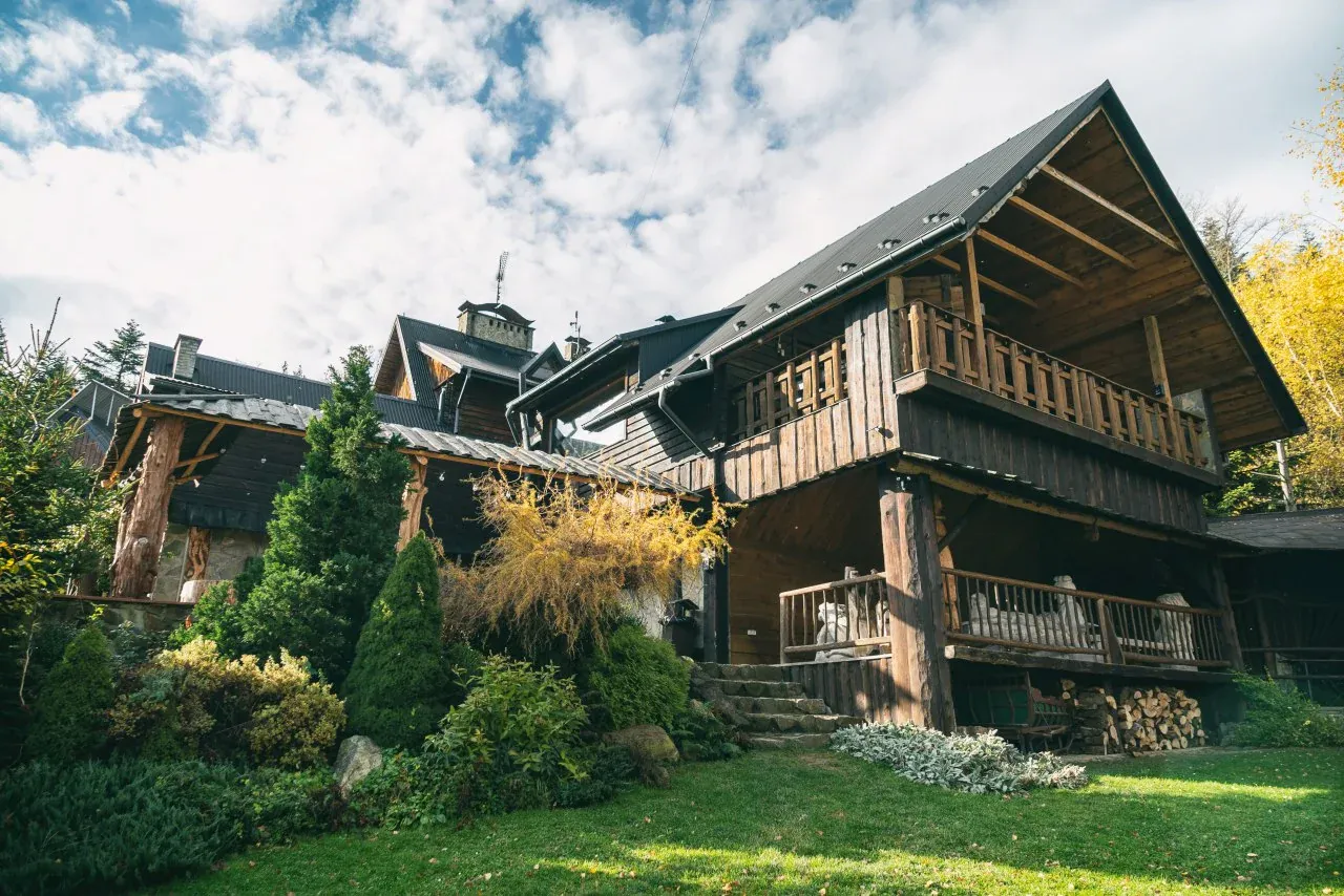 Drewniana bac&oacute;wka Biały Jeleń w Iwkowej, otoczona zielenią i jesiennymi drzewami, z widocznym tarasem i stertą drewna.