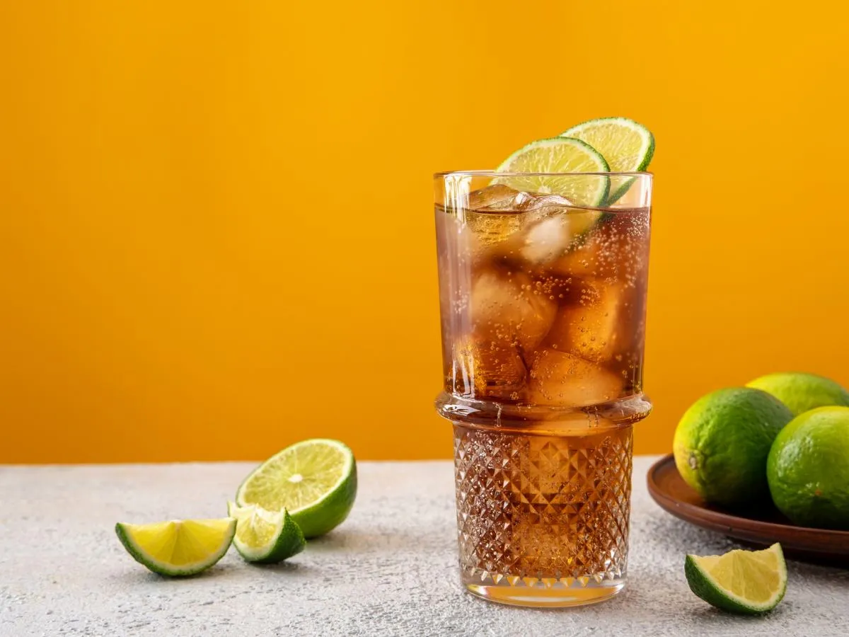 Orzeźwiający Cuba Libre, czyli rum z colą i limonką, z kostkami lodu i plasterkami cytrusów na tle pomarańczowej ściany.