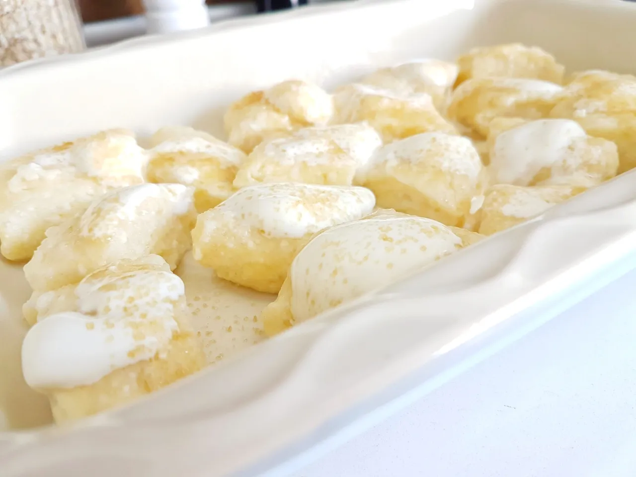 Gotowe do pieczenia pierogi leniwe w naczyniu, polane śmietaną i posypane cukrem. Idealne na szybki deser.