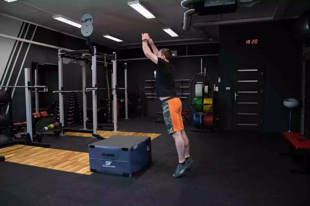 Mężczyzna wykonuje skok na skrzynię, ćwicząc jak trenować crossfit. W tle siłownia z wyposażeniem.