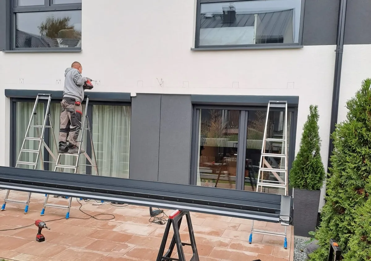 Mężczyzna na drabinie montuje element pergoli. Widać narzędzia i przygotowania do pracy, jak zamontować pergole w ogrodzie.