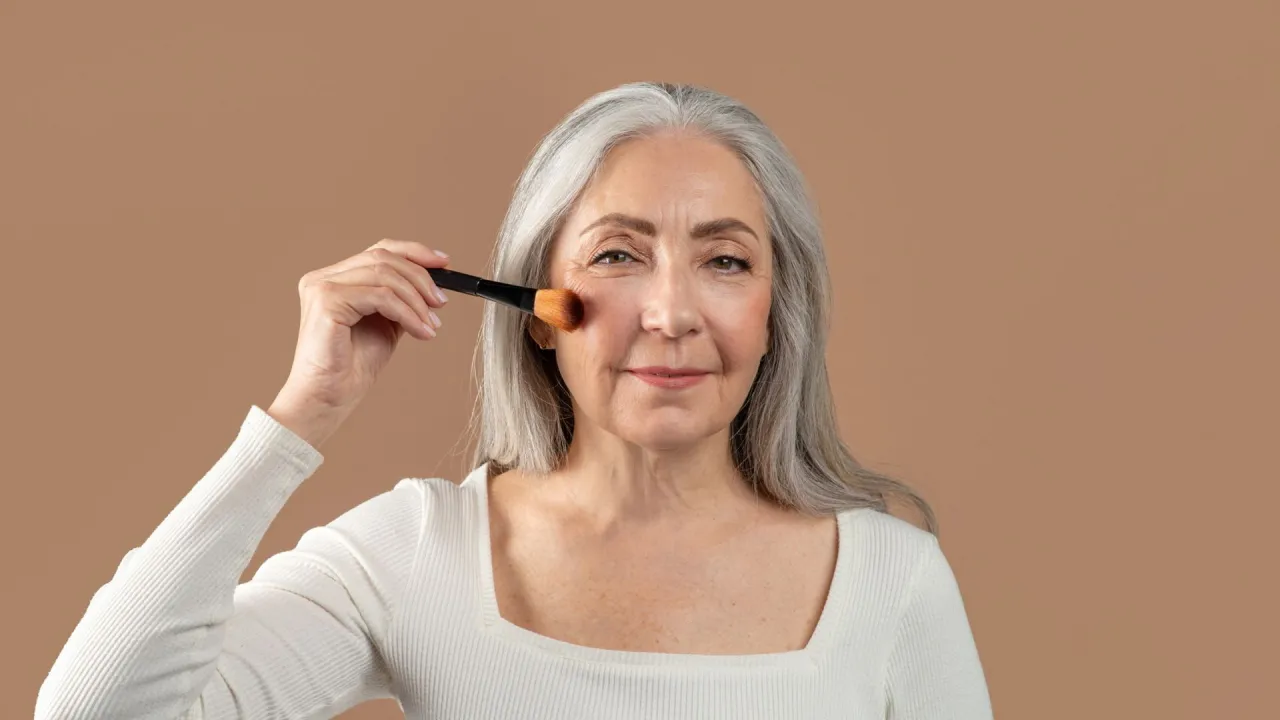 Elegancka kobieta po 50-tce z siwymi włosami nakłada r&oacute;ż pędzlem. Szuka idealnego podkładu po 50-tce.