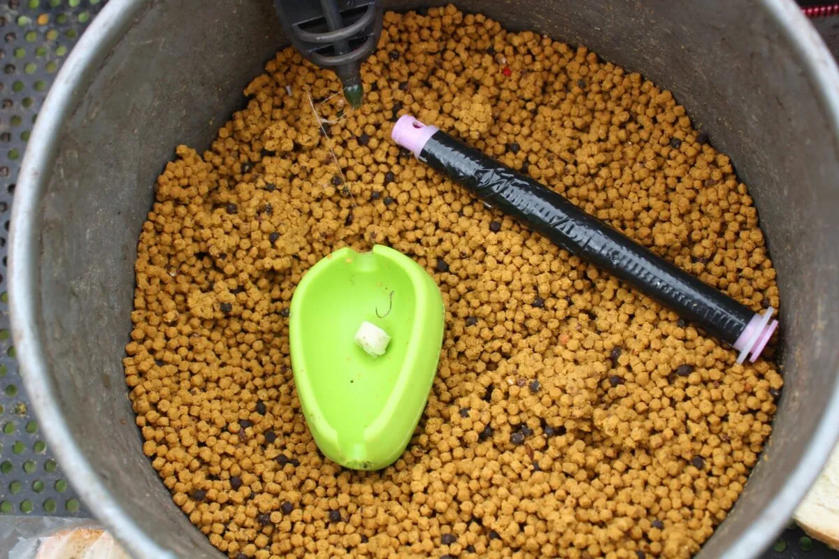 Zielona ł&oacute;dka z haczykiem i kawałkiem chleba, gotowa do zarzucenia. W wiadrze zanęta, jak założyć chleb na haczyk.