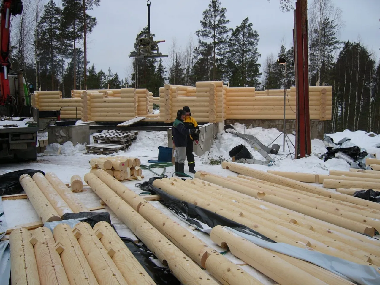 Budowa domu z bali w zimowej scenerii. Widać postęp prac, stosy bali i pracownik&oacute;w. Ile trwa budowa domu z bali? Zależy od wielu czynnik&oacute;w.