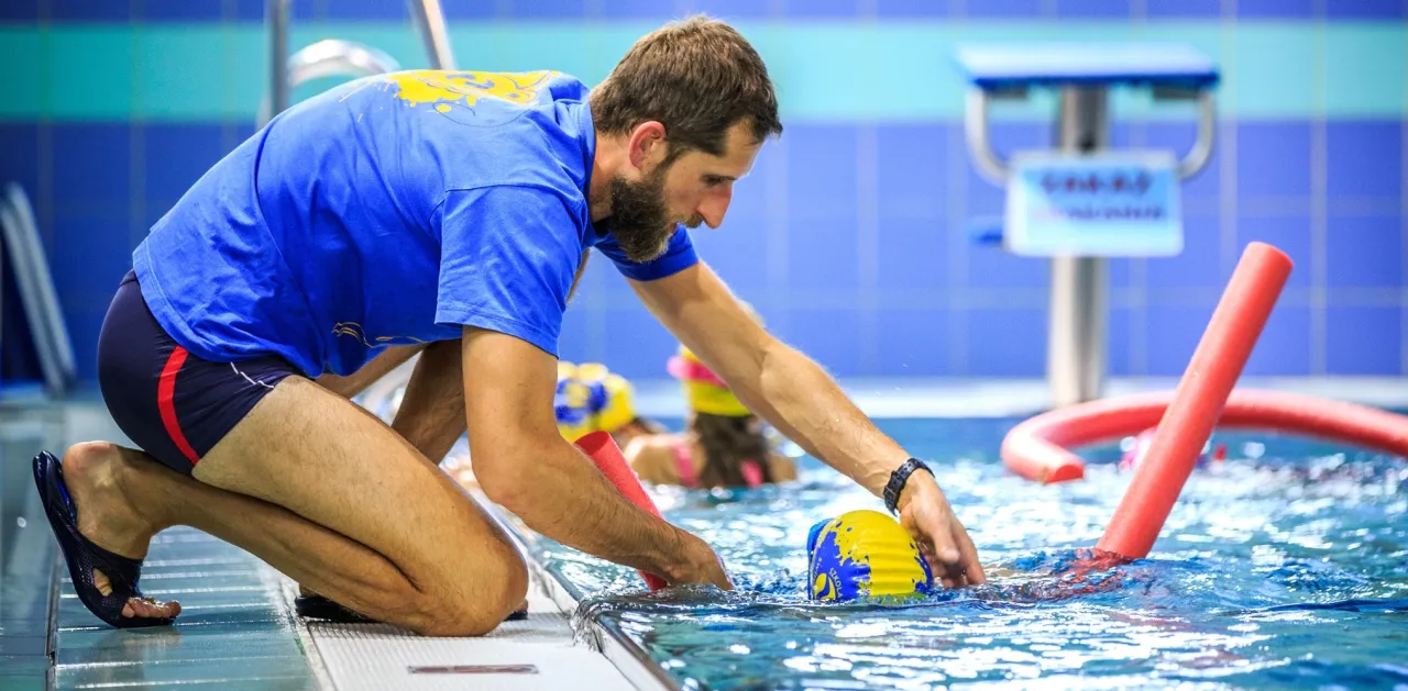 Instruktor pomaga osobie w nauce pływania w basenie. W tle widać inne osoby i makarony pływackie. To świetny przykład, jak nauczyć się pływać w starszym wieku.