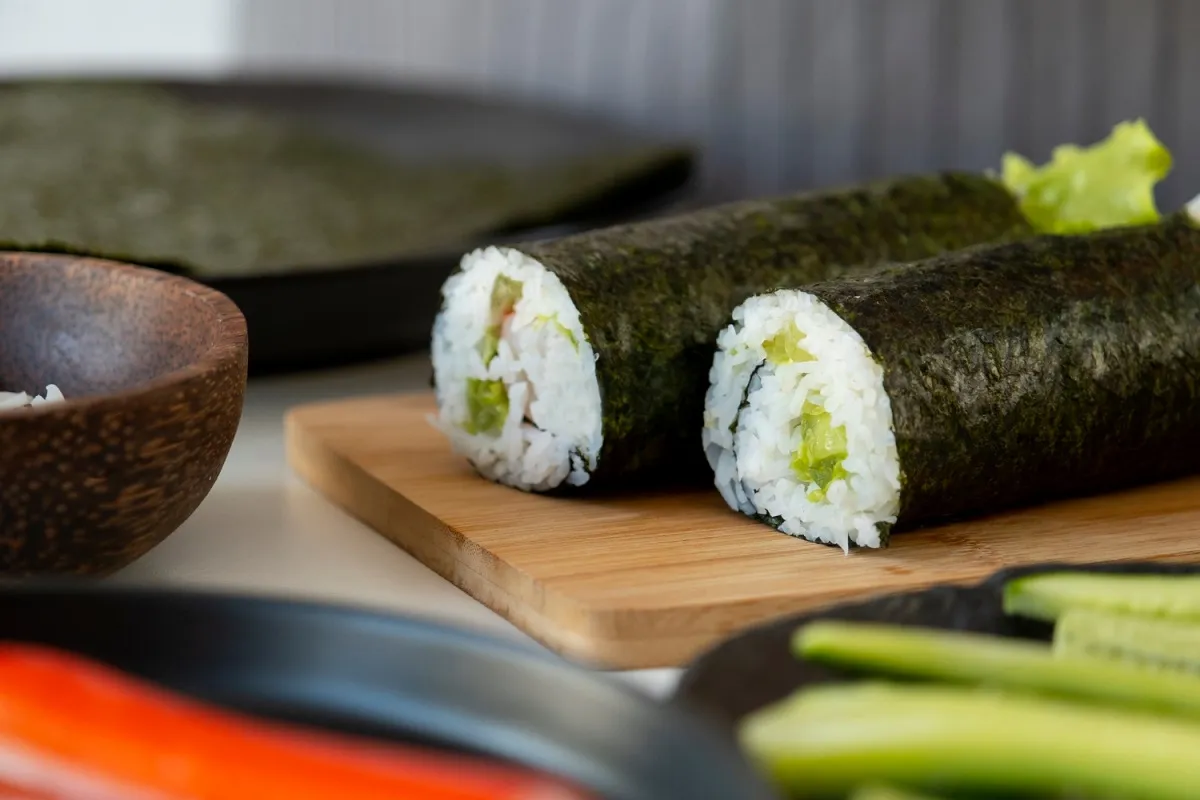 Dwa rolki sushi z ogórkiem i ryżem, zawinięte w nori, leżą na desce. Obok miseczka z ryżem i pokrojony ogórek.