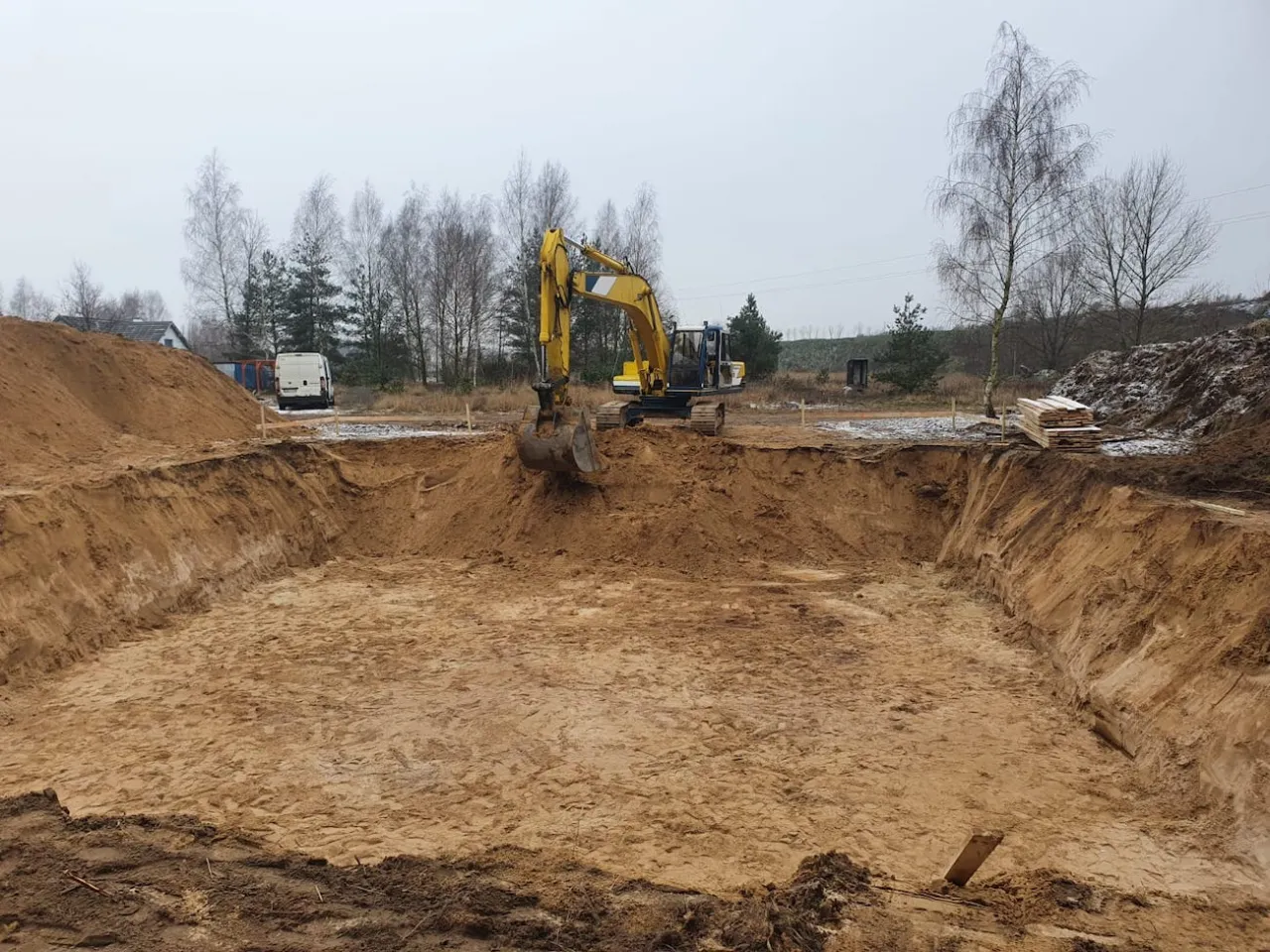 Koparka pracuje przy wykopie pod fundament. W tle widać drzewa i budynki.