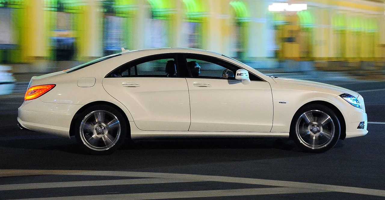 Biały Mercedes CLS 63 AMG pędzi nocą przez miasto, rozmazując światła budynków.